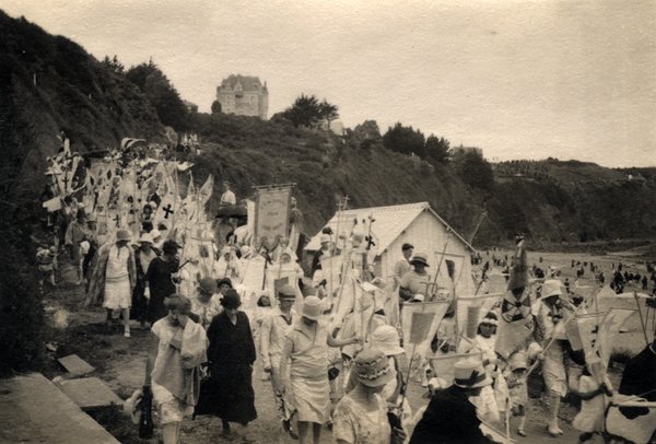 Longue procession religieuse le long d'une plage, château sur la falaise en arrière-plan. Photo ancienne sépia.ᴬᴵ