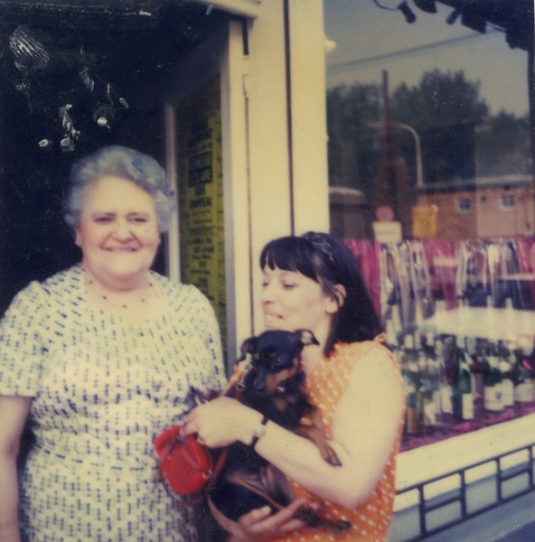 Femme âgée souriante dans l’embrasure d’un magasin côte à côte avec une femme brune un chien dans les bras.