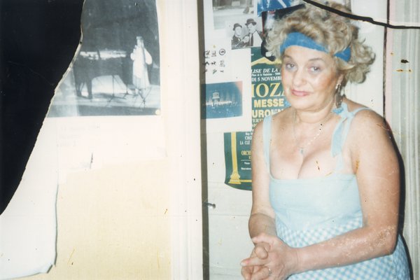 Femme mûre blonde avec un bandeau bleu assise en robe décolletée, affiches en arrière-plan. Image déchirée.