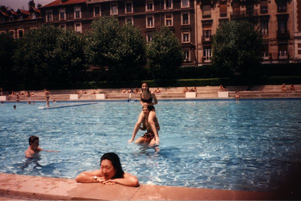 Piscine extérieure avec immeubles en arrière-plan, baigneurs dans l'eau.ᴬᴵ