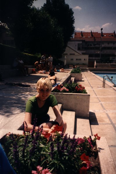 Garçon blond assis près de parterres de fleurs au bord d'une piscine.ᴬᴵ