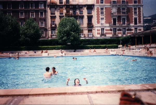 Femme faisant un signe de la main dans une piscine, immeubles en arrière-plan.ᴬᴵ
