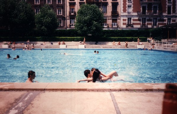 Un couple s'enlace dans une piscine, immeubles en arrière-plan.ᴬᴵ