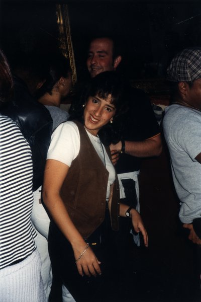 Jeune femme souriante à l'objectif, un homme derrière elle, soirée en intérieur.ᴬᴵ