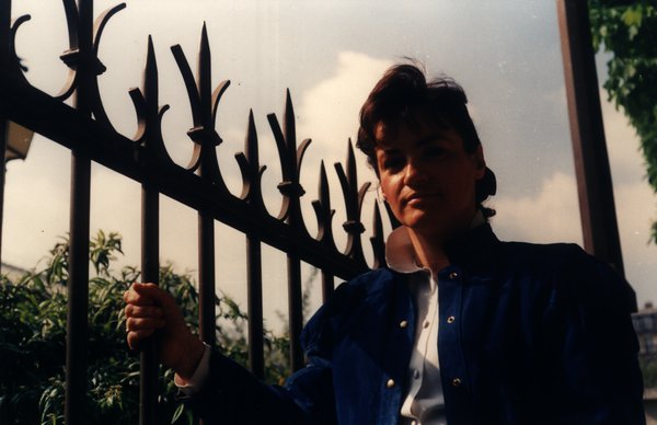 Femme en veste bleue à contre-jour près d'une grille en fer forgé.ᴬᴵ