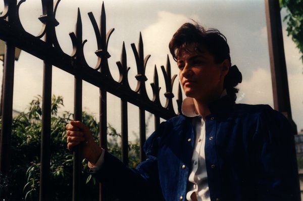 Femme en veste bleue regardant de côté près d'une grille en fer forgé.ᴬᴵ
