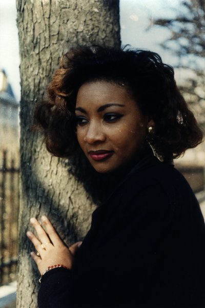 Femme aux cheveux bouclés appuyée contre un arbre, mains jointes.ᴬᴵ