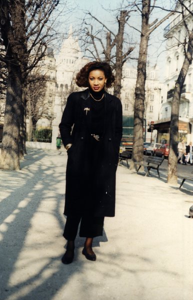 Femme en long manteau noir debout dans une rue, basilique du Sacré-Cœur en arrière-plan.ᴬᴵ