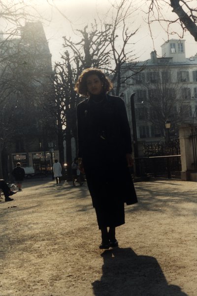Femme en manteau noir marchant sur une place, passants en arrière-plan.ᴬᴵ