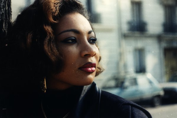 Portrait d'une femme aux cheveux bouclés appuyée contre un poteau, dans une rue.ᴬᴵ