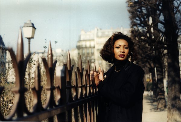 Femme aux cheveux bouclés près d'une grille en fer, rue parisienne.ᴬᴵ