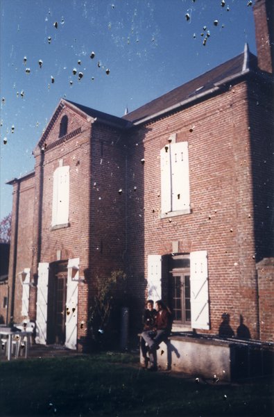 Façade de maison en brique et volets blancs, deux personnes assises sur un parapet en avant-plan.