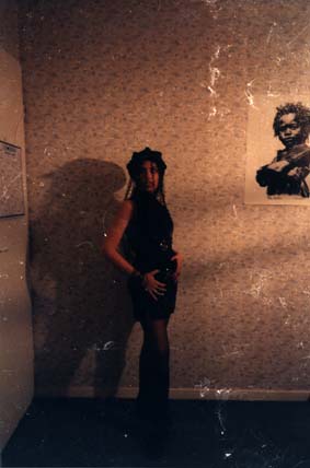Jeune fille  à casquette et mini jupe noire pose devant un mur recouvert de papier à fleurs, affiche en arrière-plan.