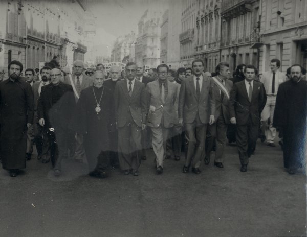 Rassemblement de manifestants avec en première ligne un prêtre et des élus municipaux. Noir et blanc.