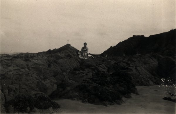 Personne assise sur des rochers au bord de la mer, croix en arrière-plan. Noir et blanc.ᴬᴵ