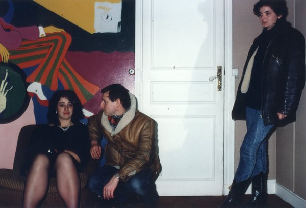Homme en blouson en peau de mouton et femme assis dans un appartement, fresque murale colorée en arrière-plan.