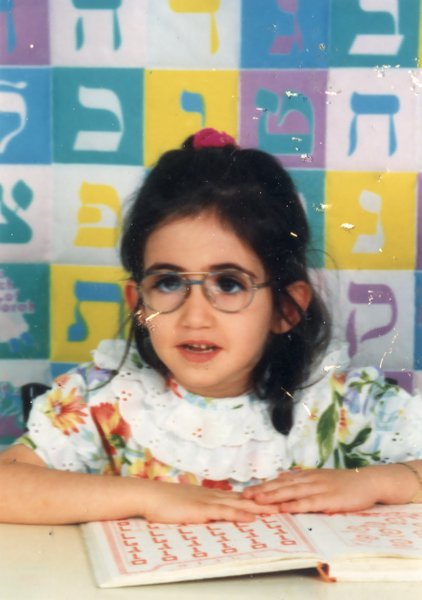 Little brunette girl with glasses in front of a table, colored Hebrew letters in the background.