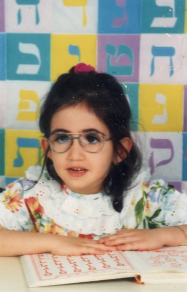 Petite fille brune à lunette devant une table, lettres hébraïques  de couleur en arrière-plan.