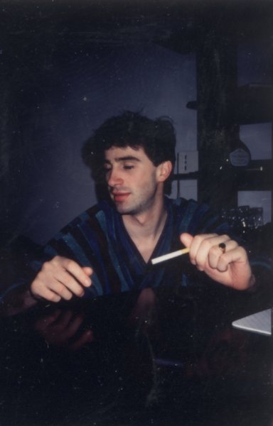 Homme brun en peignoir bleu assis avec une cigarette dans la main.