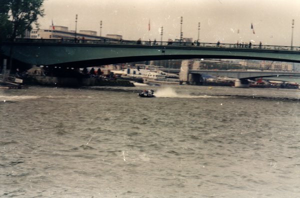 Bateau de course passant sous un pont avec des spectateurs et des drapeaux.ᴬᴵ