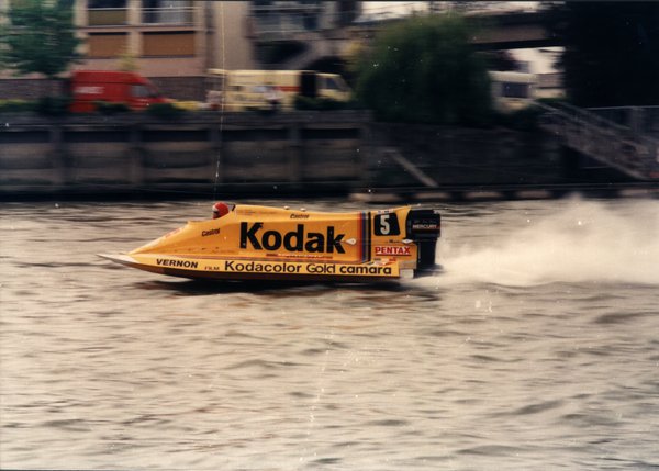 Bateau de course jaune sponsorisé par Kodak, en pleine course sur une rivière.ᴬᴵ