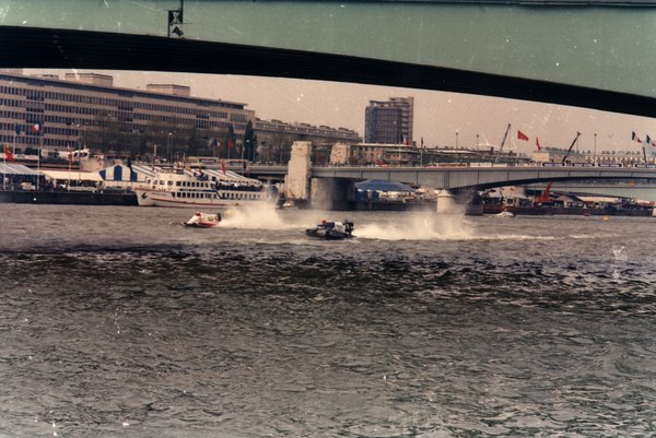 Deux bateaux de course sous un pont en ville, immeubles en arrière-plan.ᴬᴵ
