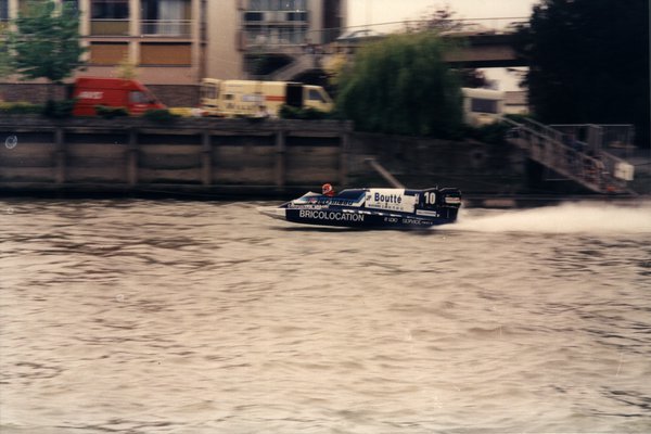 Bateau de course Boutté numéro 10 longeant des quais avec des camionnettes garées.ᴬᴵ
