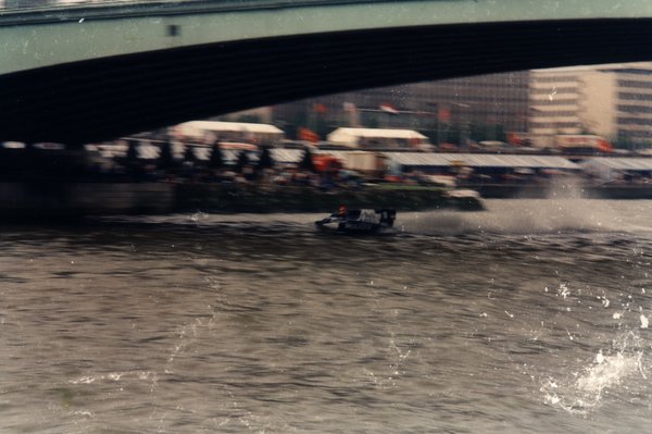 Bateau de course sous un pont, tentes et drapeaux sur les quais environnants.ᴬᴵ