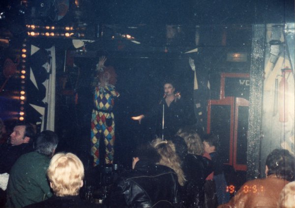 Transformiste en Edith Piaf accompagné d’un homme déguisé en arlequin sur une scène face à un public.