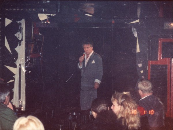 Chanteur en costume gris avec un micro sur une scène face à un public.