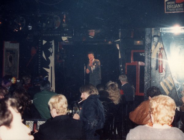 Chanteur en costume gris avec un micro sur une scène face à un public.