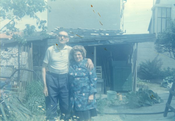 Homme et femme âgés à lunettes  debout accolés devant une cabane de jardin.
