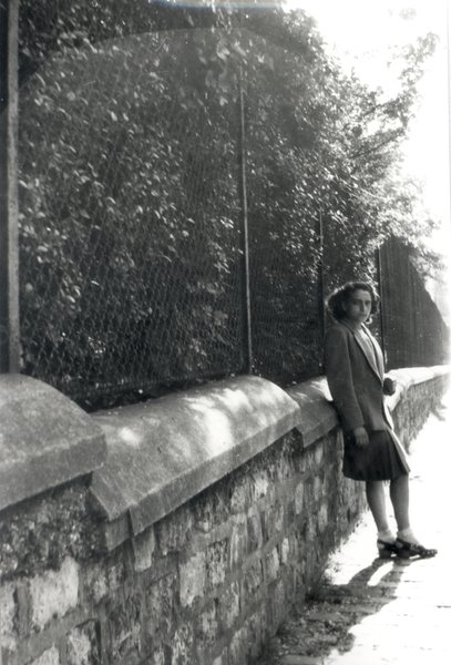 Jeune femme brune en jupe plissée adossée à un mur grillagé. Noir et blanc.