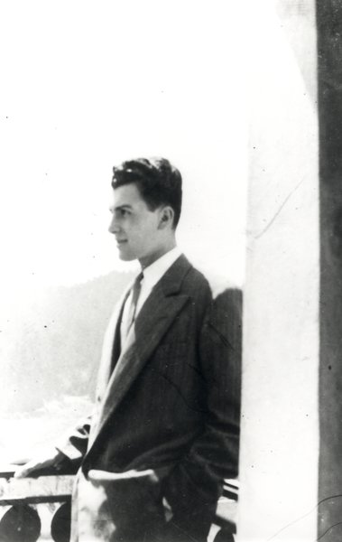 Jeune homme brun souriant debout de profil sur un balcon.