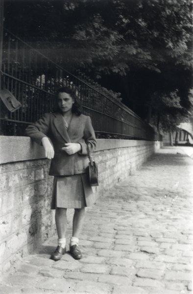 Femme en jupe avec un sac à main accoudé au muret grillagé d’un square. Noir et blanc.