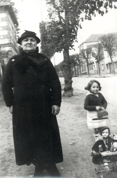 Femme âgée en habit et chapeau noir accompagnée d’une fillette avec une poupée dans une poussette.