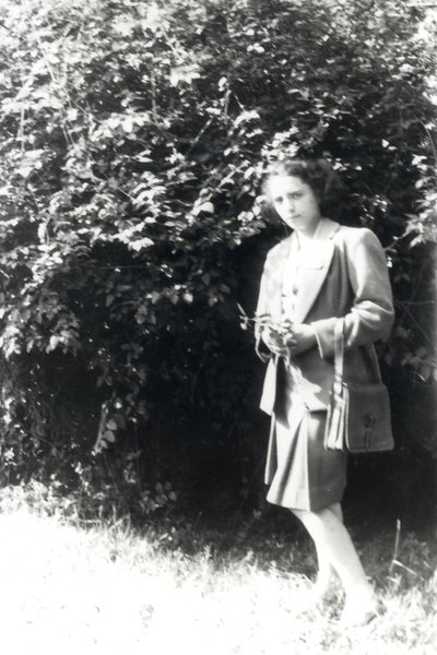 Femme avec une jupe plissée, une fleur à la main, debout devant un buisson. Noir et blanc.