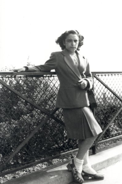 Jeune femme en jupe plissée et socquettes, accoudée au parapet d’un pont métallique. Noir et blanc.