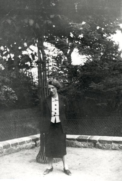 Jeune femme en jupe et veste sombre debout devant un arbre. Noir et blanc.