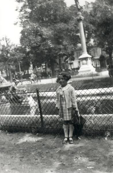 Petite fille en robe debout avec un parapluie devant bassin et fontaine dans un square. Noir et blanc.